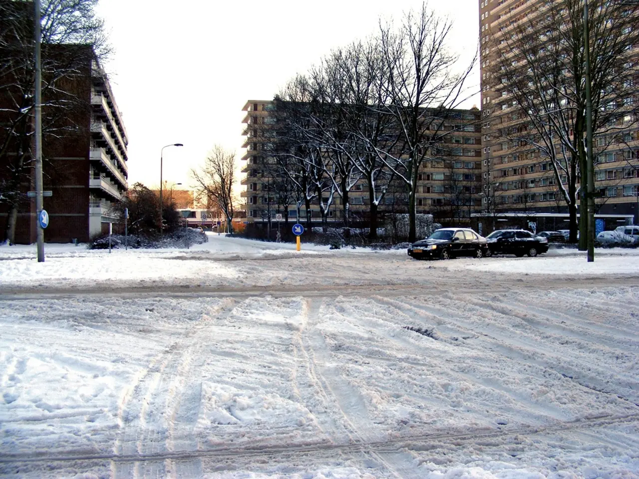In this picture we can see vehicles on the snowy road with poles, trees and buildings on either...