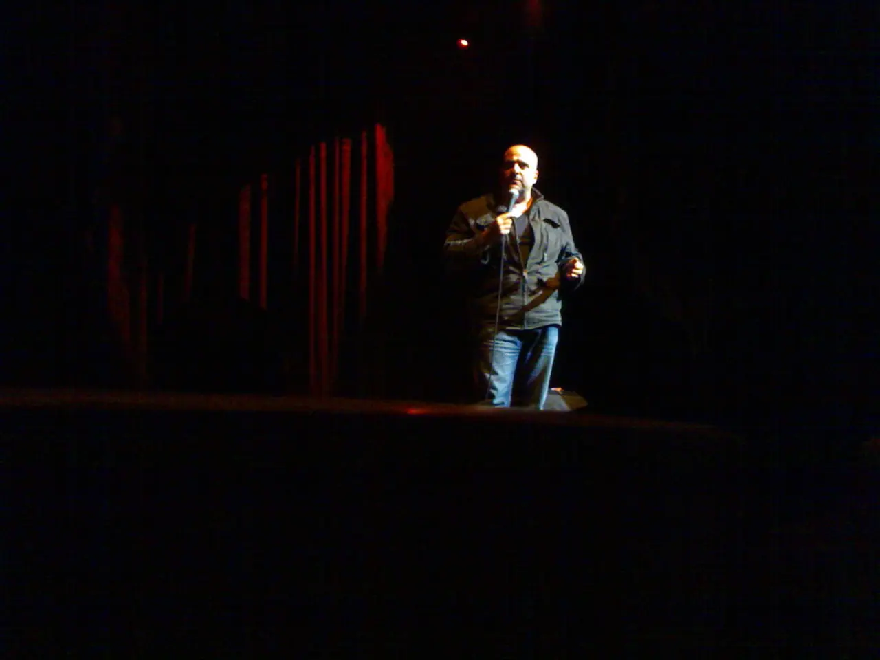 In this picture I can see a man speaking into microphone, at the top there is a light. In the...