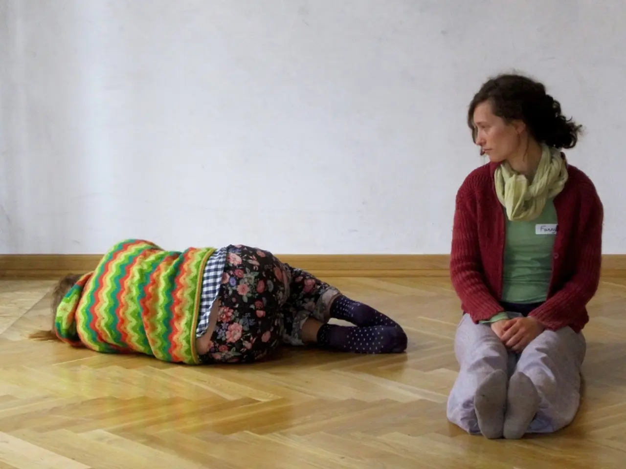 In this image there are two people, a wooden floor and a wall. Among them one person is lying on...