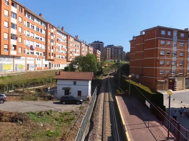 In this image we can see sky, buildings, fences, trees, ground, motor vehicles on the road, railway...