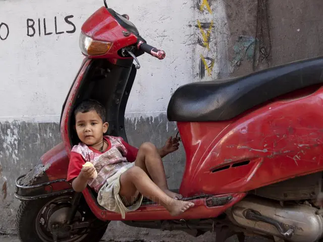 There is a scooter. On that a boy is sitting and holding something in the hand. And there is a wall...